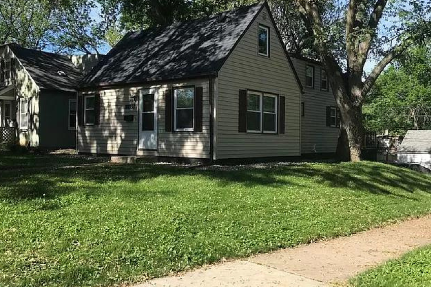 A quaint Cape Cod-style home with beige siding and white window frames, nestled among mature trees in a lush green lawn, located at 2201 S Euclid Ave, Sioux Falls, SD.